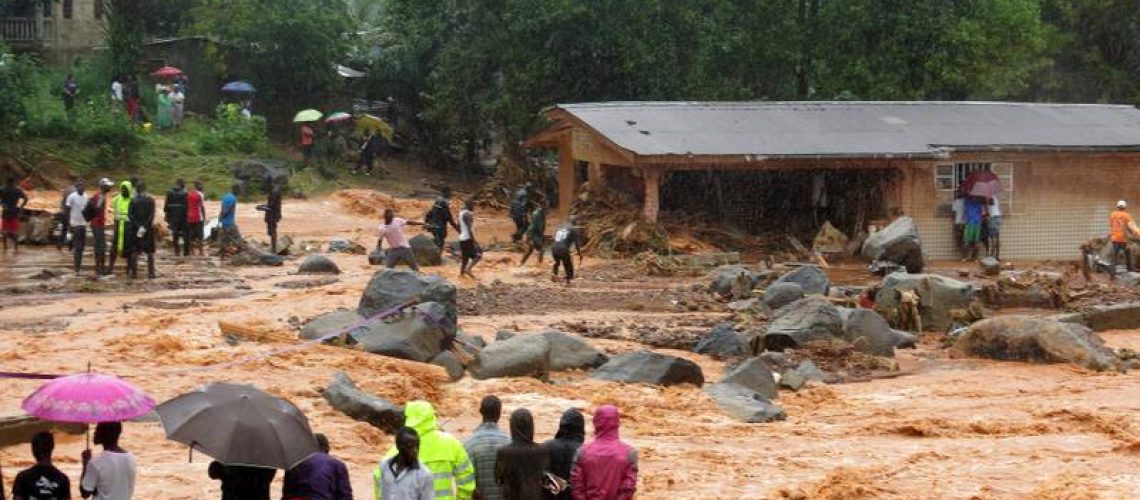 Bystanders look on as floodwaters rage past a damaged building in an area of Freetown on August 14, 2017, after landslides struck the capital of the west African state of Sierra Leone.               
At least 312 people were killed and more than 2,000 left homeless when heavy flooding hit Sierra Leone's capital of Freetown, leaving morgues overflowing and residents desperately searching for loved ones. An AFP journalist at the scene saw bodies being carried away and houses submerged in two areas of the city, where roads turned into churning rivers of mud and corpses were washed up on the streets.
 / AFP PHOTO / SAIDU BAH